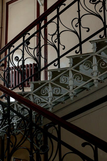 Inside a Victorian home showing a staircase with ornate black wrought iron balustrades and white wooden steps, partially visible, leading upwards. A dark wooden handrail runs along the staircase. Behind the staircase, a radiator is mounted on the wall, and a window with natural light is partially visible at the top. The environment appears immaculately maintained, with the photo capturing the structural details of the staircase and surrounding interior. This scene illustrates the type of historic architecture that may require careful handling during a home relocation or furniture transport process, as guided by companies like Man with Van Chalk Farm, to avoid staircase damage during moving and packing activities.