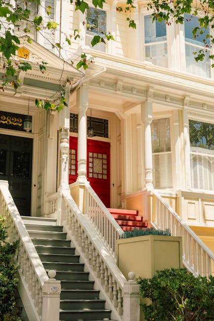 High-resolution image showing the exterior of a Victorian-style townhouse in Chalk Farm during daytime. The property features a white-painted façade with decorative architectural mouldings, large sash windows with white frames, and an ornate front porch supported by white columns. A bright red front door with glass paneling is accessible via a flight of grey-painted wooden stairs with white balustrades, leading from a small front yard with greenery and shrubs. Adjacent to the stairs, climbing plants with green leaves partially obscure the upper windows and front facade. Natural sunlight illuminates the scene, emphasizing the historic charm of the house. The image, associated with house removals and packing and moving services by Man with Van Chalk Farm, captures the careful staging involved in home relocation and furniture transport processes, with the stairs and façade illustrating common challenges in avoiding staircase damage during house removals.