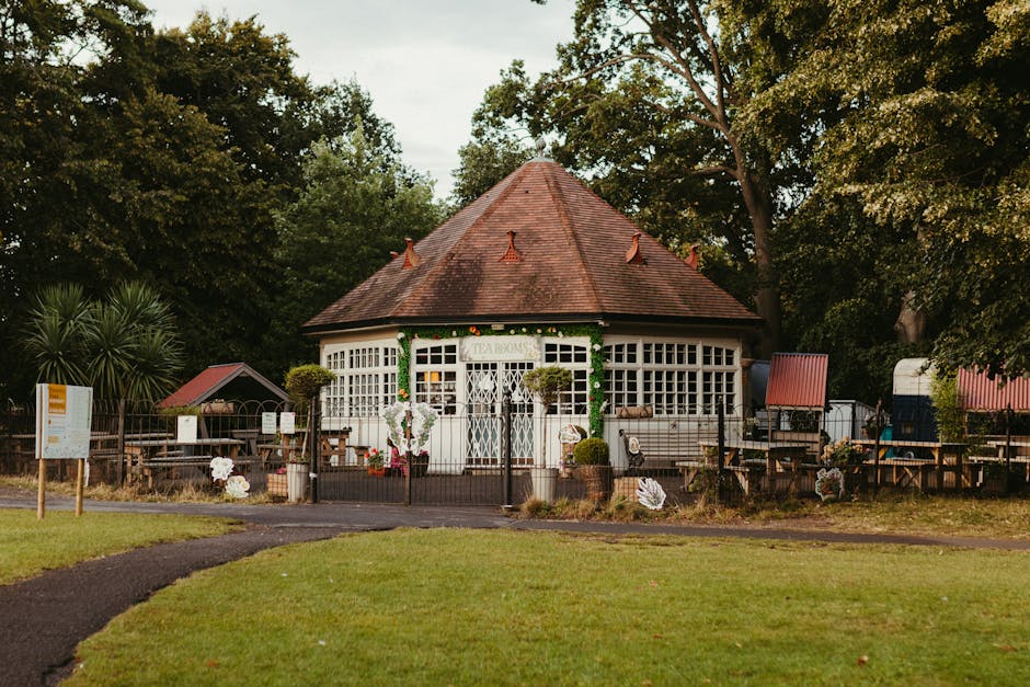 A small, circular pagoda-style building with a red-tiled, conical roof and white wooden framing, situated within a lush green park environment. The structure features multiple large, evenly spaced windows with white grid panes, and a central entrance with white lattice double doors. Green garland decorations are draped around the upper part of the building, and two potted plants flank the doorway. Surrounding the building is a low wooden fence, with additional decorative elements such as artificial flowers attached to it. In front of the building, a paved pathway leads through a well-maintained grassy area. Tall trees with dense foliage provide shade and backdrop, creating a tranquil setting. The scene is illuminated by natural daylight, capturing the peaceful atmosphere typical of a community or park pavilion used during home relocation or community events, with the [COMPANY_NAME] offering professional removals and furniture transport services to facilitate home and furniture moves near The Roundhouse, Chalk Farm.
