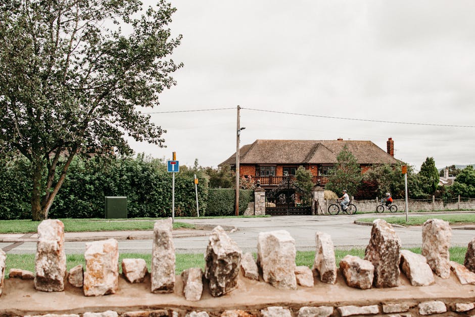 A white and orange flatbed truck loaded with wooden pallets and cardboard boxes is parked in the middle of a busy town street during daylight hours. The truck is positioned at an intersection near a row of multi-story brick and stucco buildings, which include a prominent blue-painted corner building with large windows and a sloped roof. Surrounding the truck are pedestrians walking along the sidewalk, some carrying bags or bicycles, and a few cyclists riding past. The street surface features visible lane markings and a speed limit sign indicating 20 miles per hour. Street lamps, traffic lights, and several parked bicycles are also present, with a partly cloudy sky overhead. This scene captures the logistics of a home relocation or furniture transport operation, as seen in the context of house removal services offered by [COMPANY_NAME], highlighting the loading process and urban moving environment.