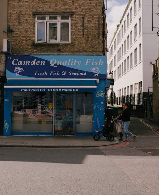 The image shows a street scene in Chalk Farm with a small fish and seafood shop called Camden Quality Fish situated on the ground floor of a brick building. The shop has a blue sign with white text, and its large glass window reveals some of the frozen fish and seafood products inside, organized on display counters. To the right of the shop, a person dressed in casual clothing is pushing a baby stroller and is standing on the pavement near the shop's entrance, possibly preparing to load or unload items related to a home relocation or moving process. The pavement is adjacent to a narrow road, and a white modern building with multiple floors is visible further down the street, suggesting an urban environment suitable for residential or commercial relocations. The scene is captured during daylight, with natural light illuminating the surroundings, and the overall setting indicates activity related to local moving or delivery services, as managed by Man with Van Chalk Farm, operating within the Camden area.