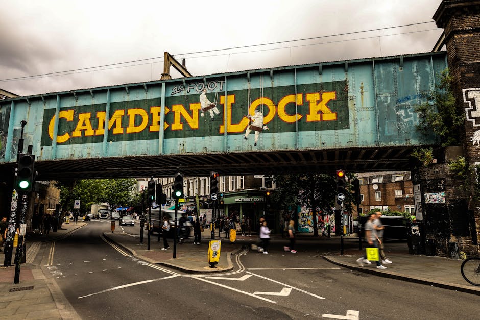 A street scene under a large, weathered railway bridge featuring the words 'CAMDEN LOCK' painted in large yellow letters on a turquoise background. The bridge demonstrates visible rust and peeling paint, with some graffiti on the right support pillar. Below, pedestrians are crossing the road, some walking and others waiting at traffic lights, which are showing green. To the sides of the street are shops, trees, and buildings, with a variety of vehicles including cars and vans on the road. In the foreground, there are bollards, road markings, and a yellow traffic cone, indicative of urban traffic management. This scene captures elements of home relocation logistics, such as the transport of furniture and boxes, with visible movement and activity suggesting a busy urban environment where professional removals, like those offered by Man with Van Chalk Farm, could assist with house moves around Camden area. The environment appears to be during daytime with overcast weather, natural lighting, and street furniture highlighting the typical cityscape backdrop for removals and moving services.