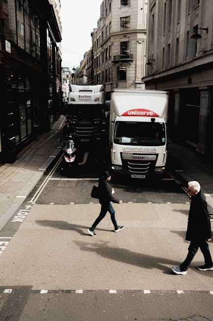 A street scene showing a white moving van with the branding 'uniserve' parked partially on the pavement and partially over the railway tracks, with the rear doors closed. The van is positioned adjacent to a narrow alleyway between tall, multi-storey buildings typical of an urban area. In front of the van, a man is loading or unloading items, though specific objects are not visible. A person with a black backpack, wearing dark clothing, walks across the pedestrian crossing near the van, while another older individual with white hair and dark clothing walks in the opposite direction. The residential buildings feature large windows, some with fire escapes, and the street is lined with typical cityscape architecture. The scene captures the typical logistics involved in house removals or home relocation, with moving equipment and positioning on the street as part of the loading process, supporting professional removals services offered by Man with Van Chalk Farm.