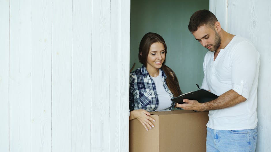 A man and a woman are positioned near a doorway during a home relocation process, with the man standing and holding a clipboard or tablet, and the woman smiling behind a large cardboard moving box. Th