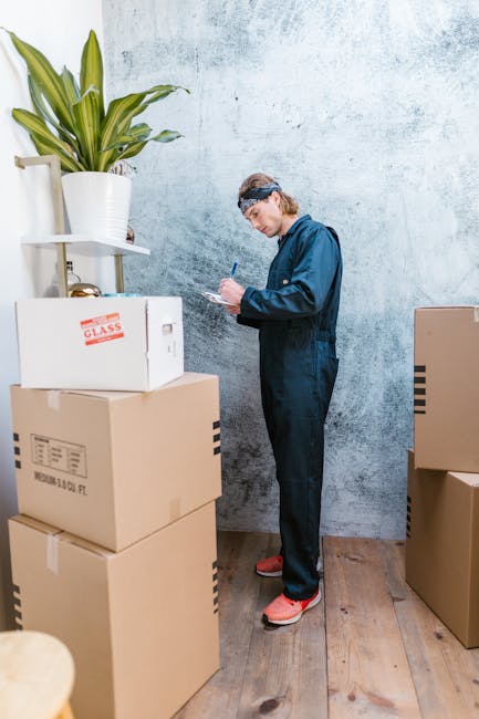 A man and a woman are positioned near a doorway during a home relocation process, with the man standing and holding a clipboard or tablet, and the woman smiling behind a large cardboard moving box. Th