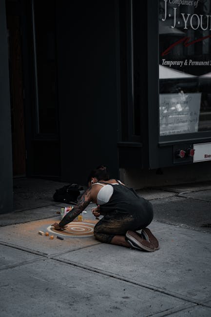 A person sitting on their knees on a sidewalk outside a building, engaged in a street art activity or chalk drawing on the pavement. The individual is wearing a white sleeveless top and dark pants, with tattoos visible on their arms. Nearby, there are multiple chalk pieces and possibly some small containers or bottles. Behind them is a dark-colored building facade with a large window displaying signs and advertisements, and part of a black door frame. The pavement shows typical urban infrastructure, and the scene appears to be in a city environment. The image captures a moment of creative activity or street entertainment outside a commercial establishment, with natural lighting and a focus on the person's artistic process, relevant to house removals and packing processes by Man with Van Chalk Farm.