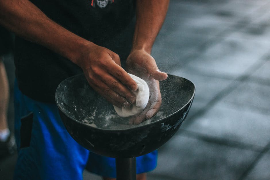 A person wearing a dark t-shirt and blue shorts is holding a black bowl in their left hand, while using a white cloth in their right hand to clean or dust the bowl's interior surface. The bowl is positioned at waist height and appears to be made of plastic or ceramic materials. The background is blurred but shows a faint water-like surface, suggesting an outdoor or nearby water setting. The scene indicates careful handling of household items, which is relevant to house removals and packing preparations, and the individual’s actions are part of a careful packing or cleaning process as part of a professional moving service by Man with Van Chalk Farm.