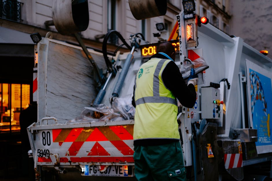 A worker wearing a high-visibility vest and blue gloves operates a waste collection vehicle parked on a residential street at dusk, with warm indoor lighting visible through a window in the background. The vehicle, branded with the logo of Man with Van Chalk Farm, has its rear open, revealing a large compartment filled with compressed waste materials, including cardboard boxes, plastic bags, and other household debris. The worker is using controls on the side of the vehicle to manage the compacting process. The vehicle's rear features red and white reflective safety markings, and the scene is illuminated by external streetlights, highlighting the loading and waste disposal activity typical of home removal and waste clearance services as part of house relocation logistics.