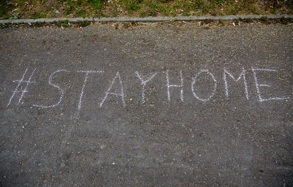 A close-up view of an asphalt pavement with white chalk writing that reads '#STAYHOME', positioned near the edge of the roadway adjacent to a strip of grass and a concrete curb. The chalk marks are clear and well-formed, situated in an outdoor environment with natural lighting. In the background, the pavement continues, indicating the location is on a street used for vehicle and pedestrian access. This image is relevant to house removals and moving services, illustrating a safe and responsible approach to home relocation, possibly reflecting message campaigns or guidance related to moving amid local street restrictions or community safety protocols, as mentioned on the Chalk Farm removals page of Man with Van Chalk Farm.
