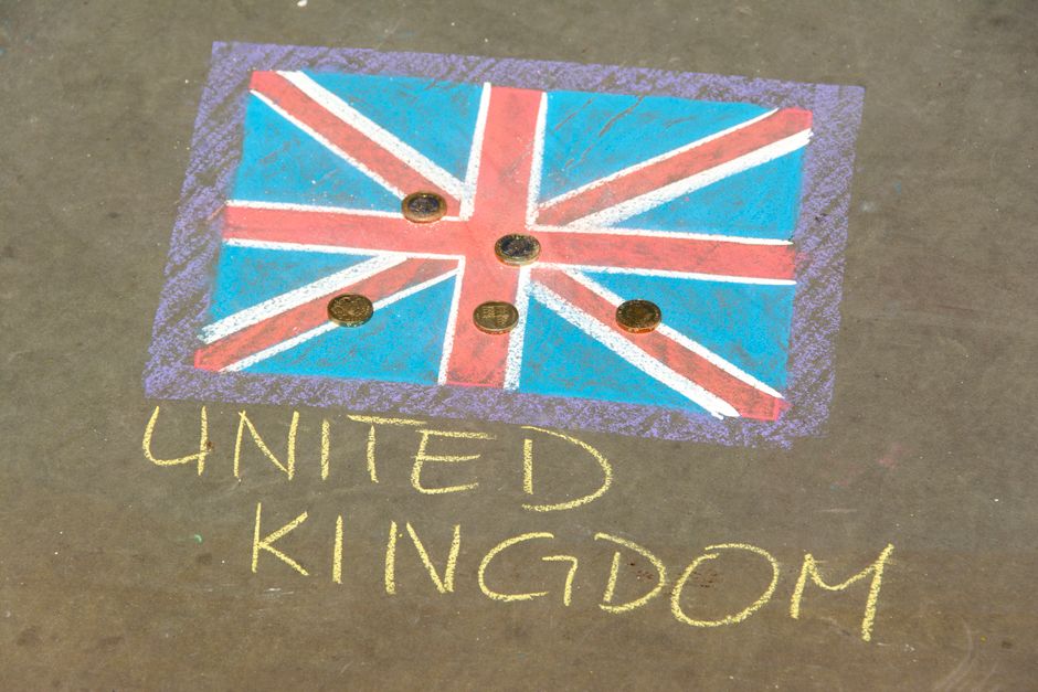 A chalk drawing on pavement depicting a stylized Union Jack flag with red, white, and blue chalk. The flag is framed by a purple border and positioned near the top of the image. Below the flag, the words 'UNITED KINGDOM' are written in yellow chalk. Surrounding the chalk artwork on the brown pavement are five British currency coins, some overlapping the flag. The scene is outdoors and well-lit, indicating daytime, and the context suggests it is related to house removals or moving services provided by Man with Van Chalk Farm, as indicated by the webpage titled 'Street Closure Rules for Chalk Farm Removals, CHALK FARM'.