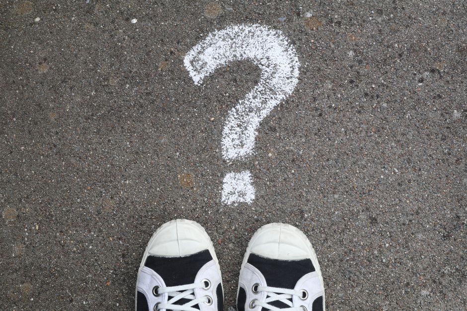 A view from above of a pair of white and black sneakers standing on a rough asphalt pavement. Directly in front of the shoes, a large white question mark is drawn on the ground with chalk. The scene a